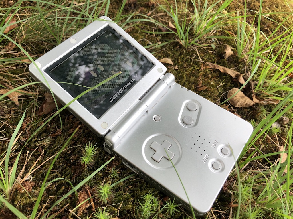 A Chickpea build running on a Game Boy Advance SP laying outside, surrounded by a variety of grasses and brown leaves, the screen partially obscured by the reflection of cloudy sunlight filtered through the trees overhead.
